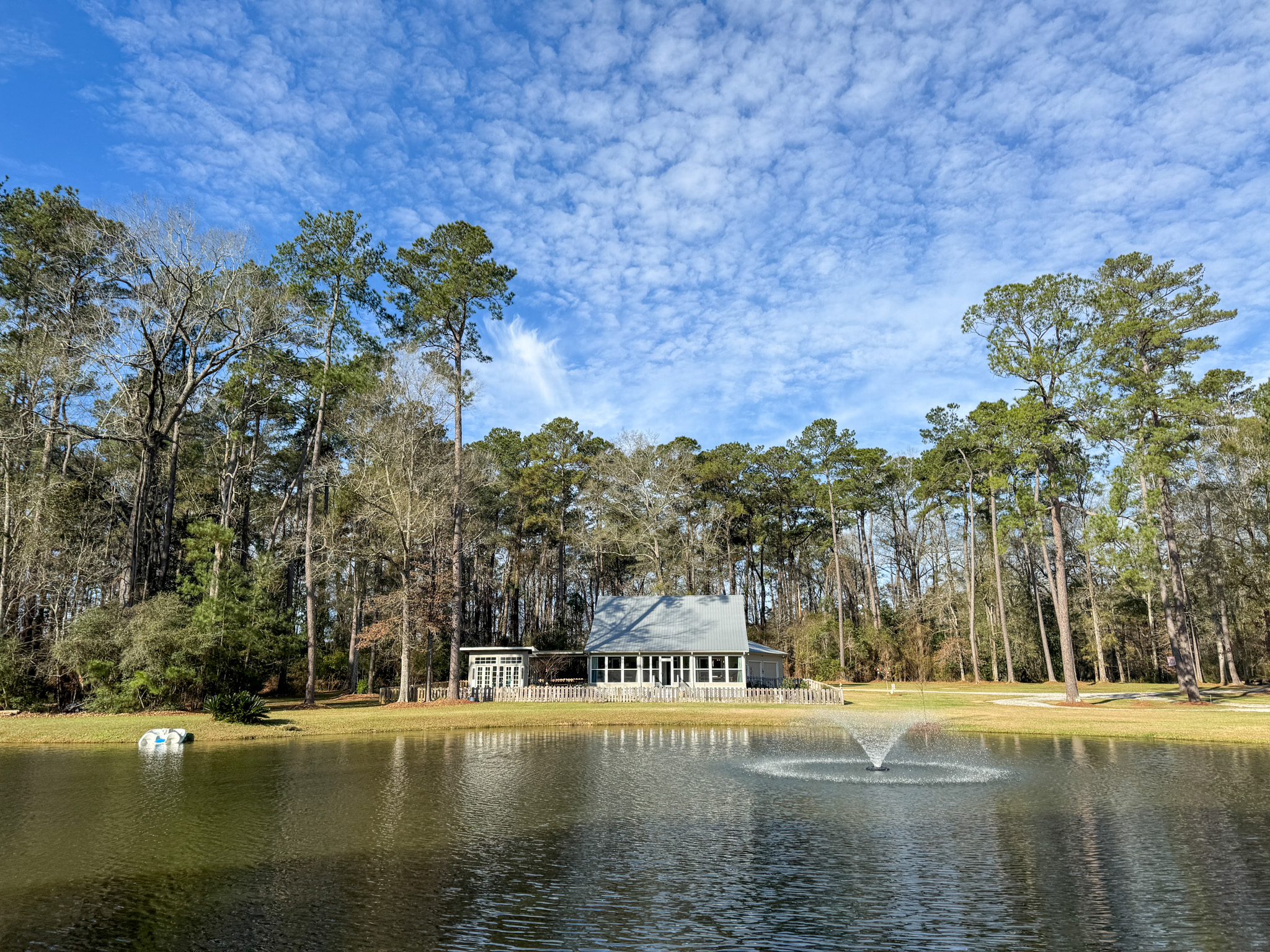 A large house with a large front porch and a small shed next to it stands across a large pond in the bright morning light.